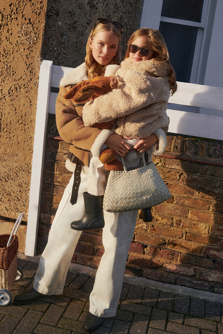 Mum and girl wear matching shearling jackets, standing outside in the sun