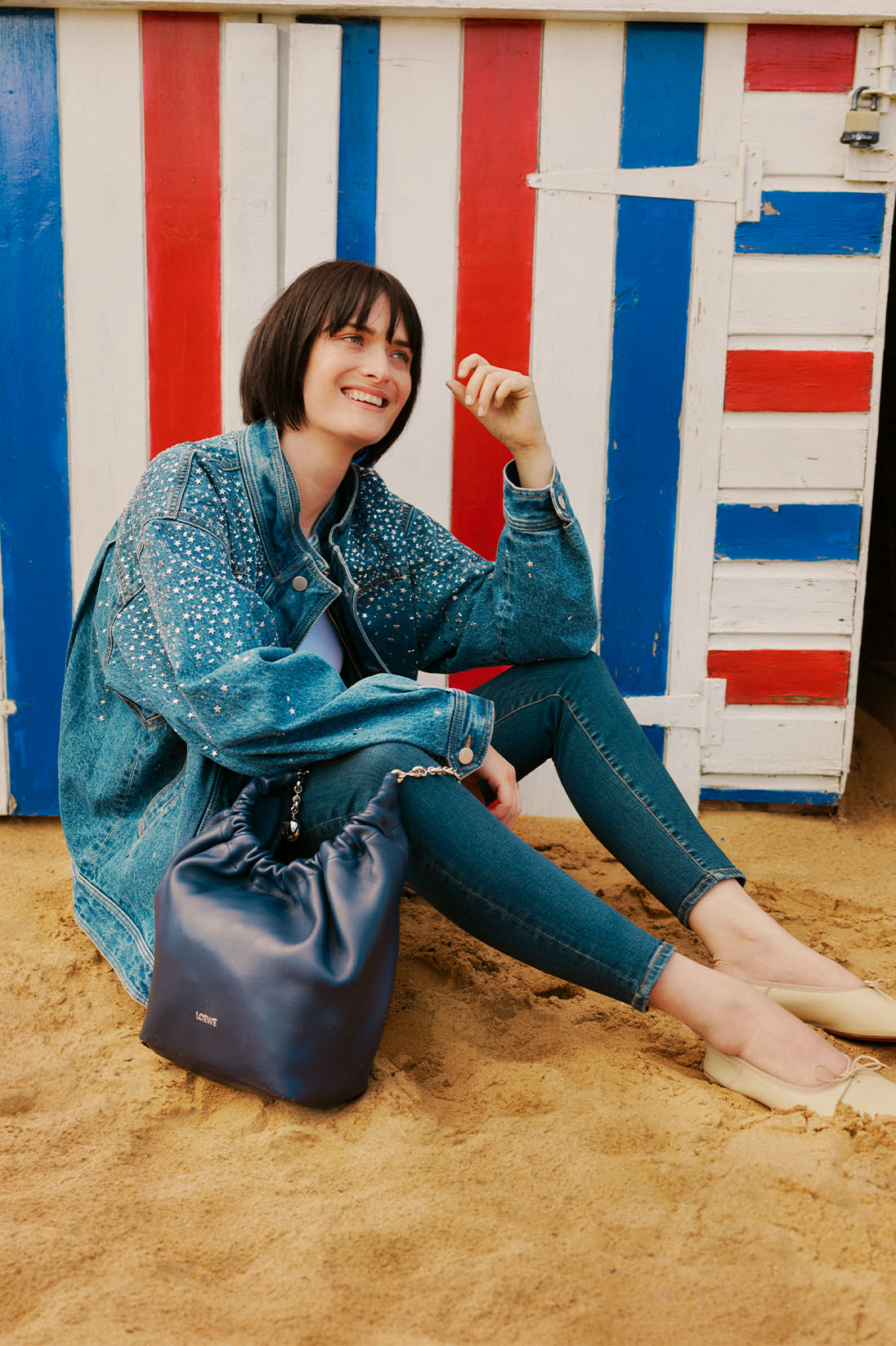 Model sits on sandy beach in full denim look including Good American skinny jeans