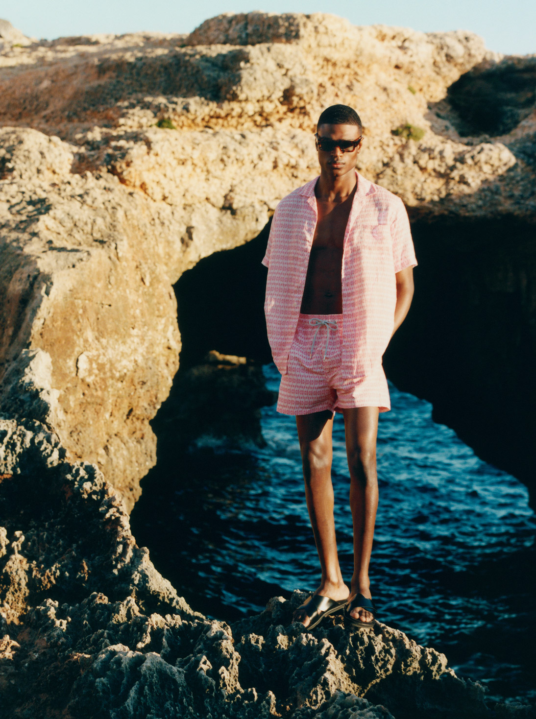 Male model stands on a rock in the sea and wears pink Love Brand & Co. shirt and shorts, Jacques Marie Mage sunglasses and TOM FORD slides