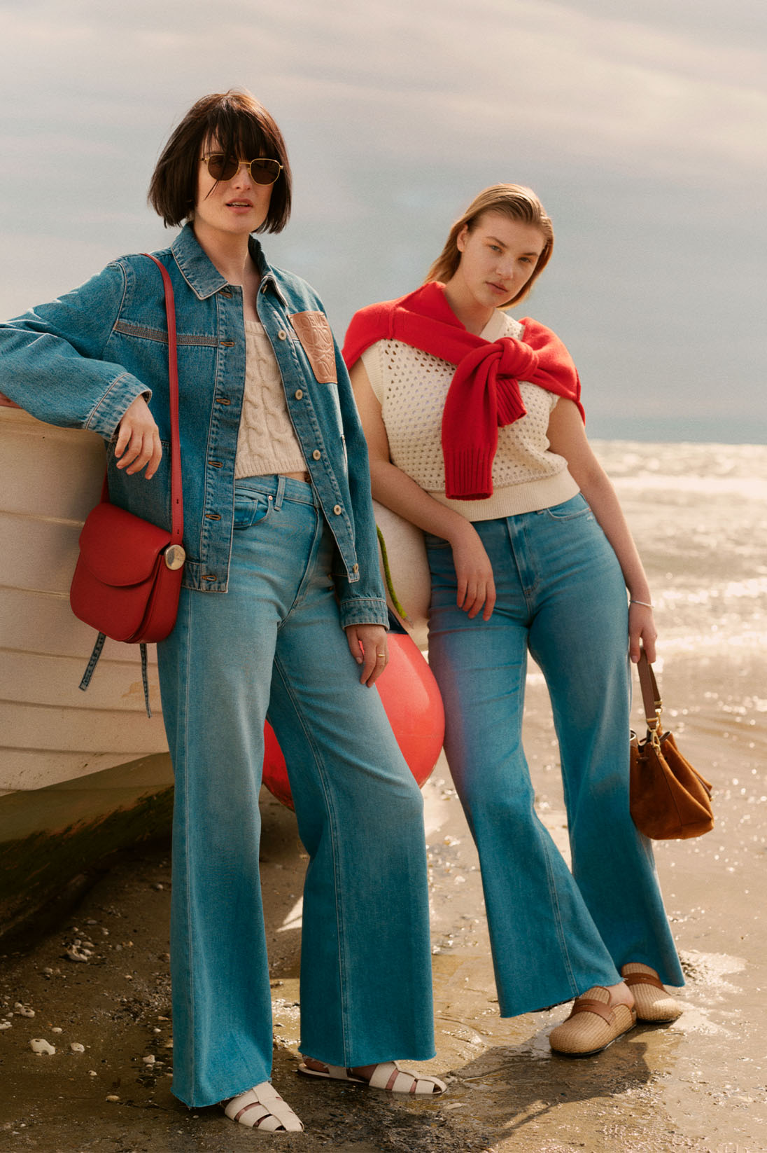 Models stand by boat on a beach wearing blue jean jacket, cream knitwear and blue Paige jeans