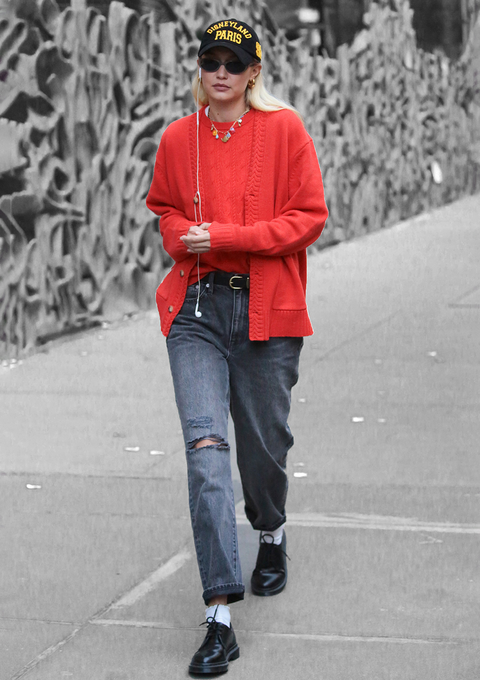 Gigi Hadid wearing a red cardigan and jumper from Guest in Residence with grey jeans, black leather shoes, a black cap and black sunglasses