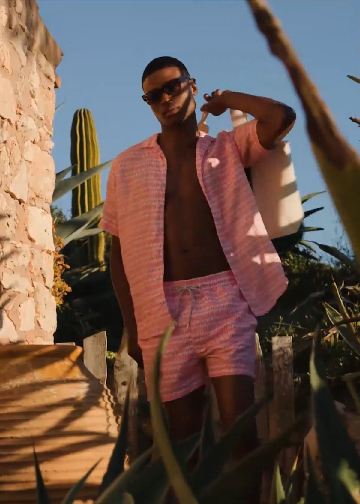 Male model walks past a wall in a sunny location and wears pink Love Brand & Co. shirt and shorts, Jacques Marie Mage sunglasses, TOM FORD slides and Vilebrequin bag