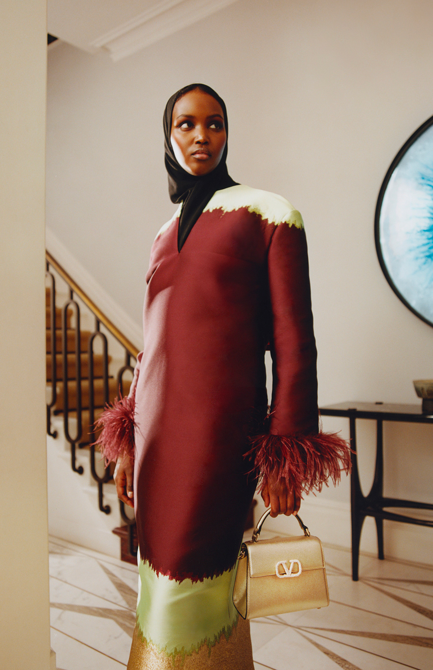 The model wears a shift-style kaftan from the brand Valentino in a deep burgundy colour, with mint-green and metallic gold accents, along with a black headscarf. The sleeves are long with burgundy feather trims, and she holds a gold top-handle bag from Valentino Garavani.