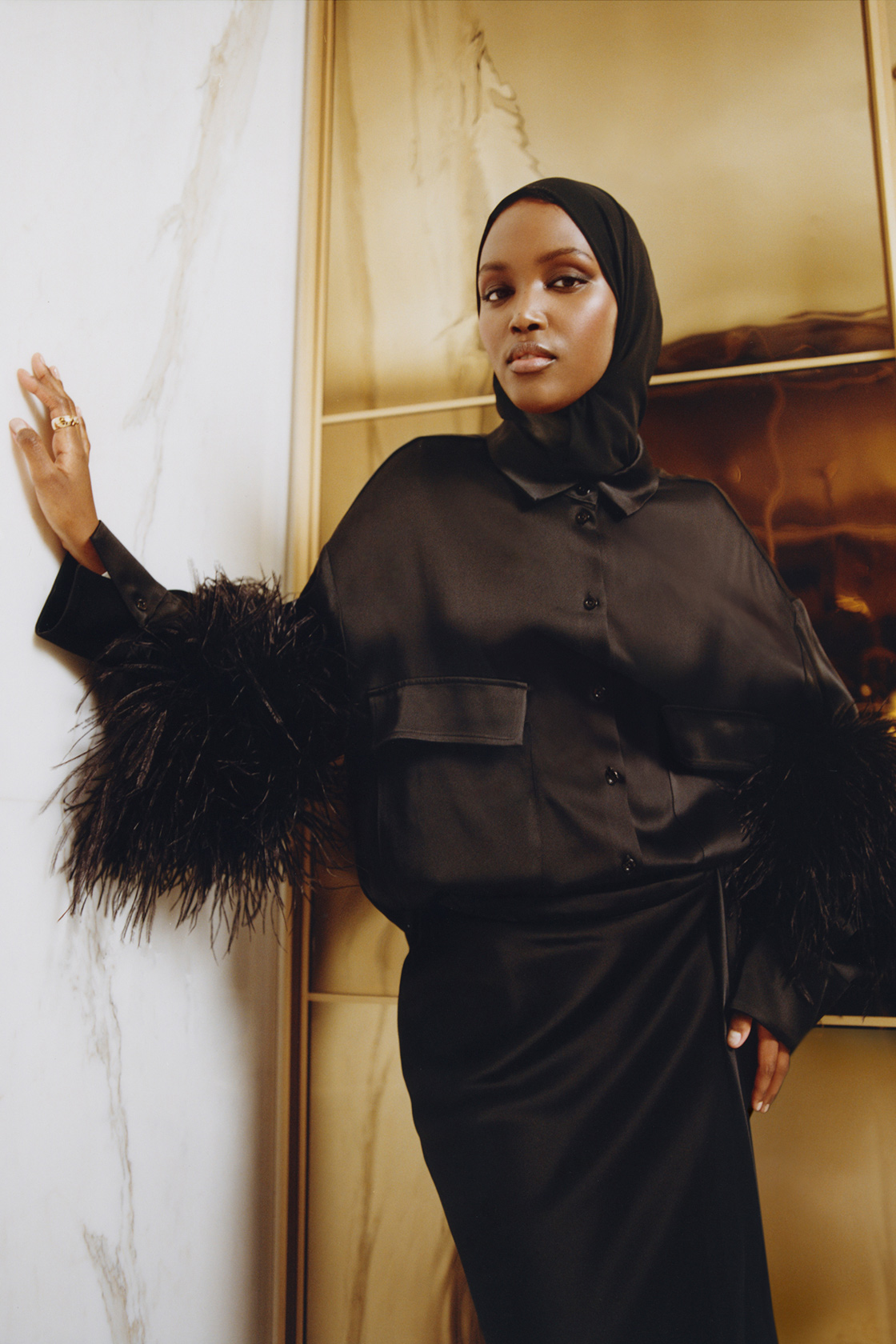 A model wears a black satin buttoned shirt with black feathers on the sleeves, with a coordinating maxi skirt, both from the brand LaPointe. She also wears a black headscarf,