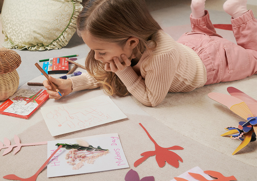 Girl sits on floor of nursery making Mother's Day cards, wearing pink jumper and trousers