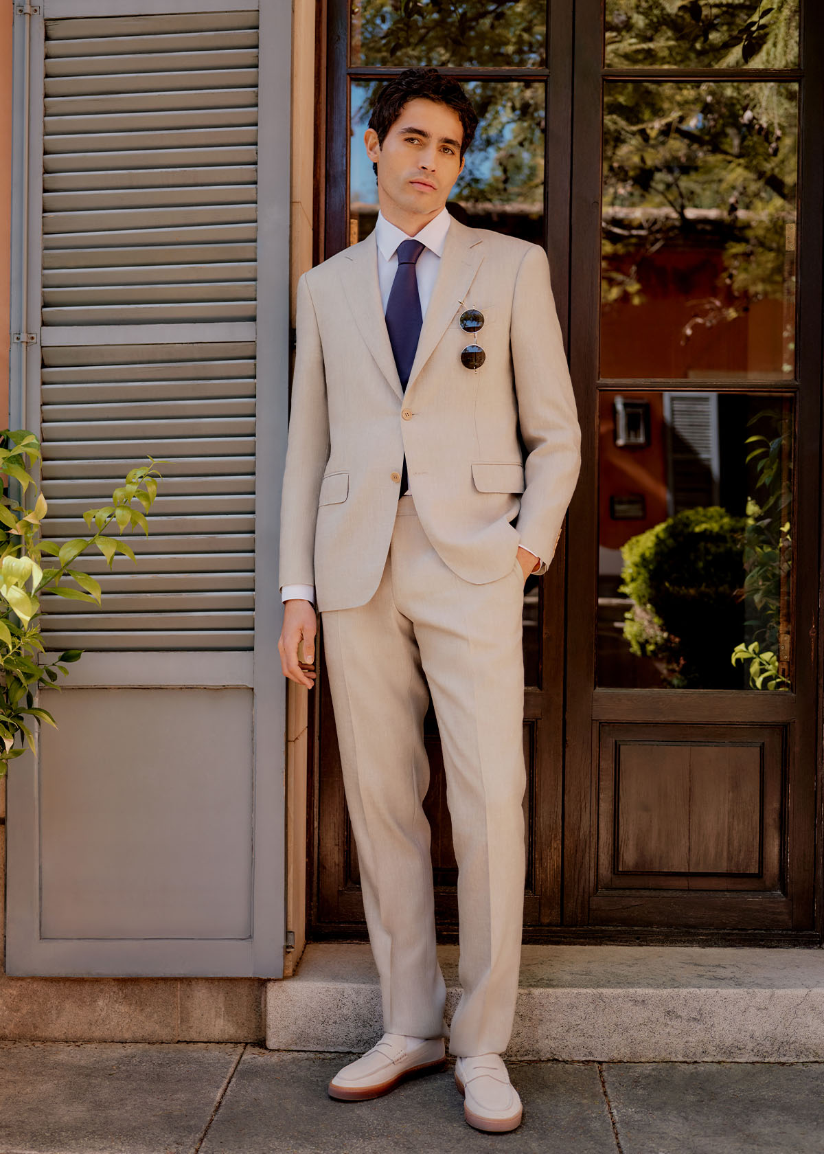 Male model wears beige Canali suit and navy tie standing in sunny location