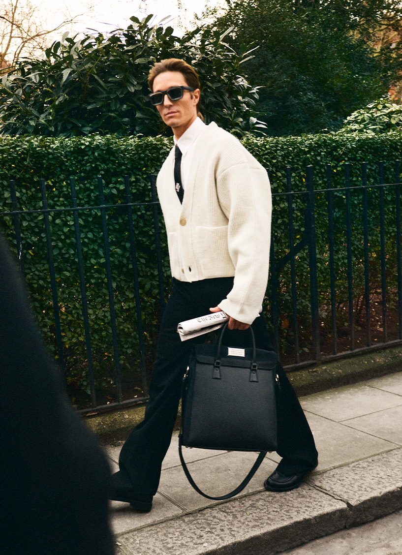 Holding a newspaper and a black bag, a male model wears a cream cardigan, shirt and black tie