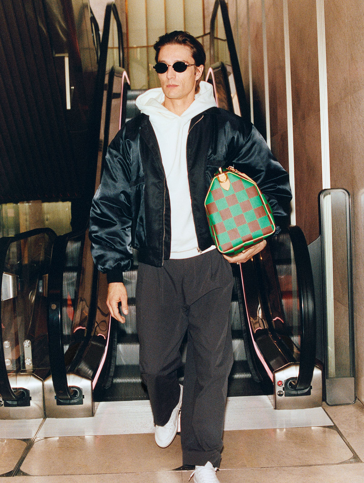 A male model walks down an escalator in Harrods wearing a black jacket, cream hoodie and green checked bag by Louis Vuitton