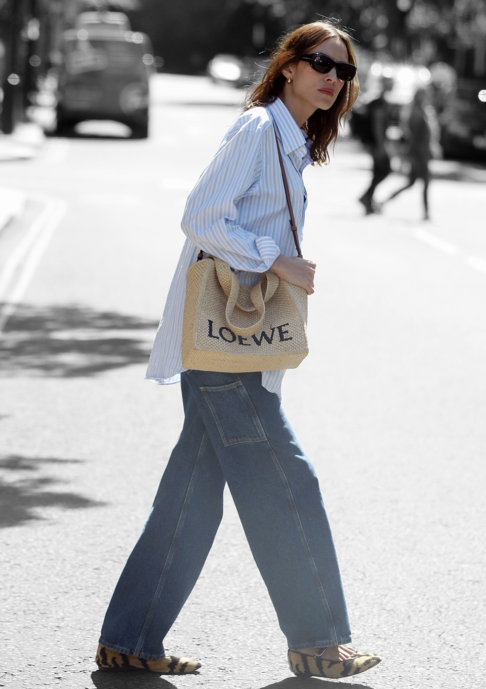 Alexa Chung crossing a street in London wearing a relaxed blue and white striped shirt with jeans, animal-print ballet flats, a Loewe woven bag and black sunglasses