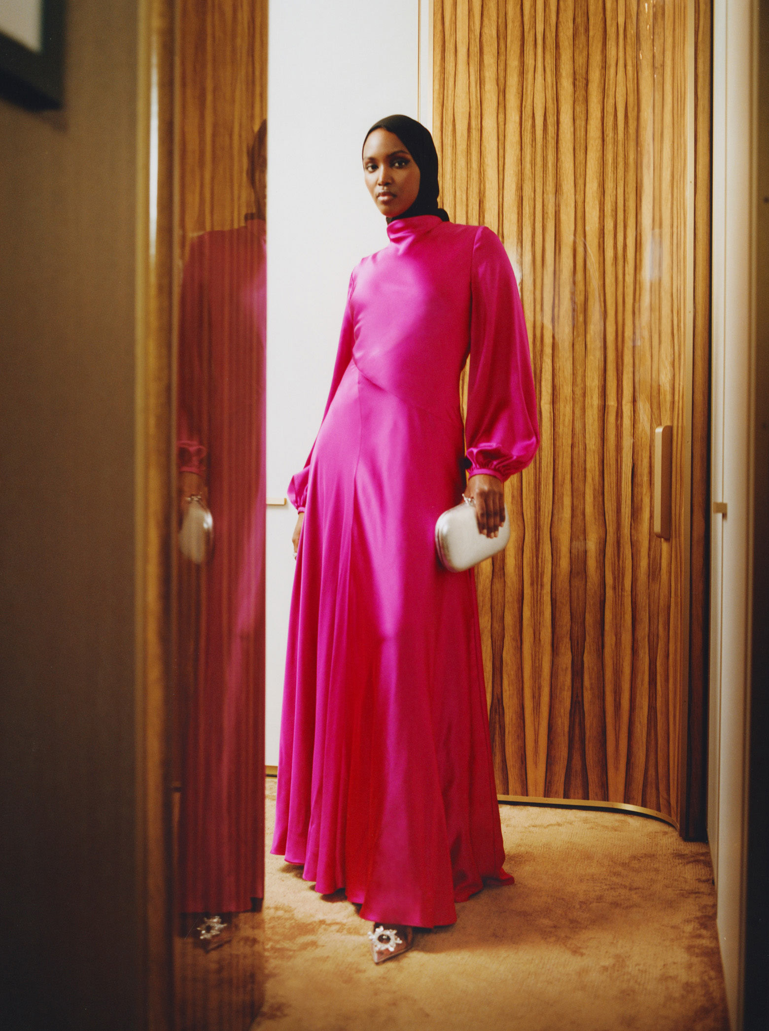 A model stands in front of a wooden door wearing a hot-pink maxi dress with long sleeves from the brand Roksanda. On her feet, she wears a pair of clear, PVC heels from the brand Amina Muaddi, with circular crystal brooches on the pointed toes, and she holds a metallic silver clutch bag from the brand Jeffrey Levinson.