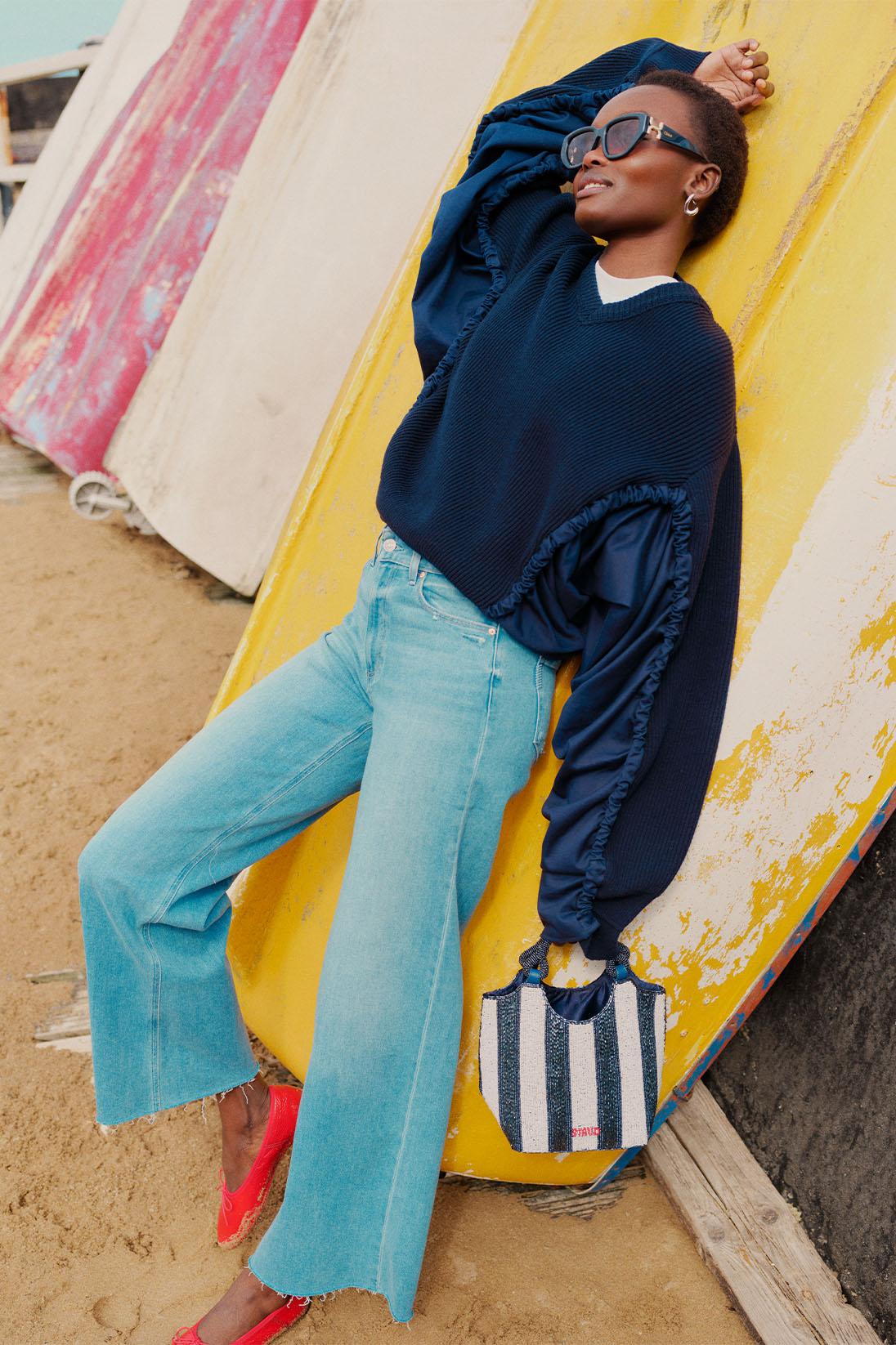 Model leans on surfboards wearing navy blue JW Anderson jumper and blue Paige denim jeans