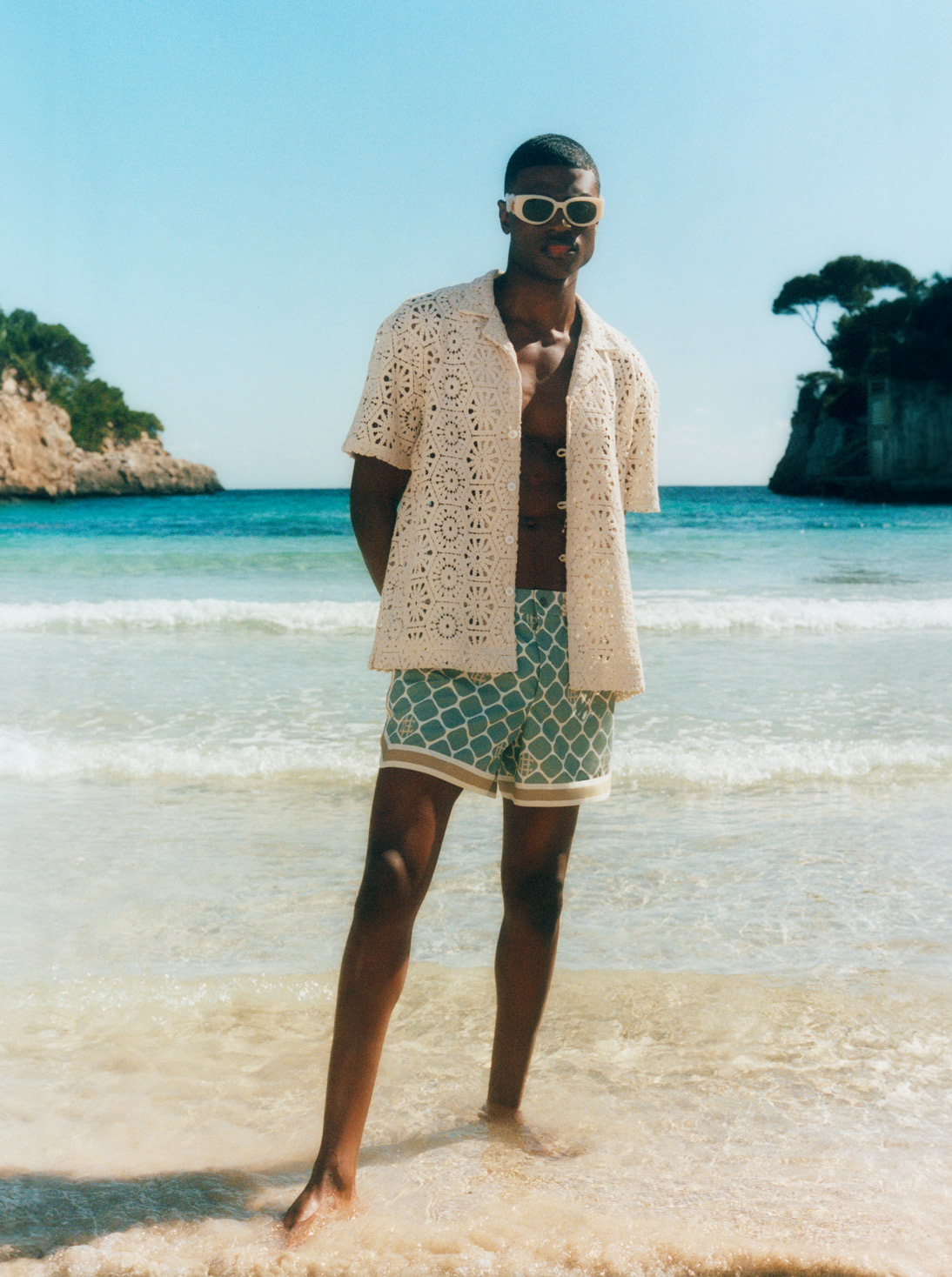 Male model stands on a beach and wears Ché shirt and swim shorts, Casablanca sunglasses, Tateossian bracelet