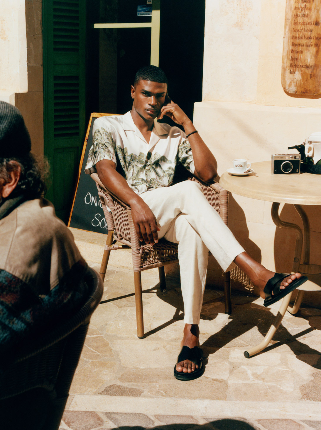 Model sits at a café in a sunny location and wears printed Eton shirt and cream Frescobol Carioca trousers