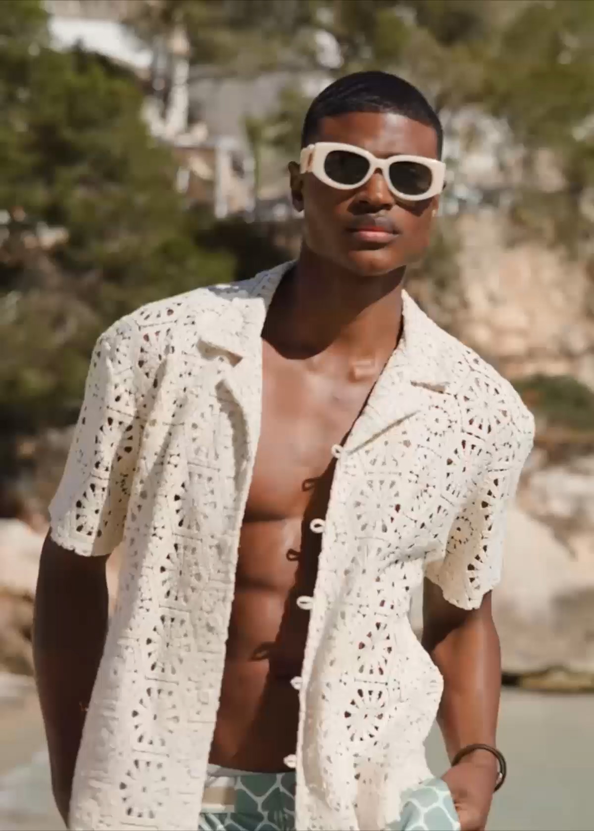 Male model stands on a beach and wears Ché shirt and swim shorts, Casablanca sunglasses, Tateossian bracelet