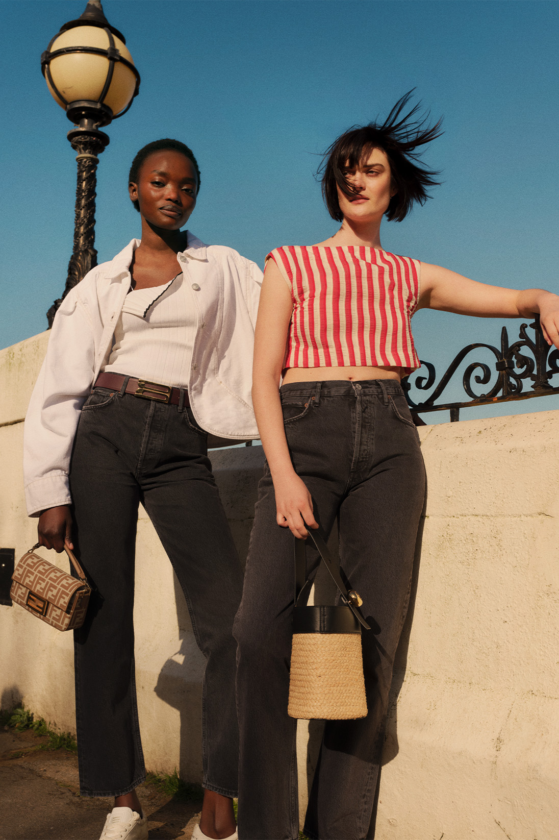 Two models stand in a sunny beach location and wear Agolde dark grey straight-leg jeans